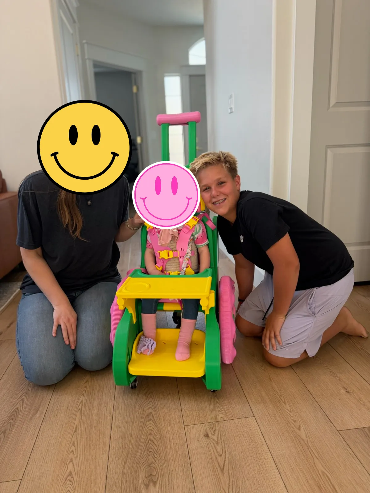 Zac kneeling beside the green, yellow, and pink 3D-printed wheelchair he built, with a toddler seated in it. The faces of the toddler and an adult are covered for privacy.